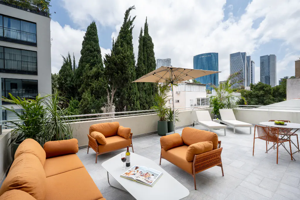 Expansive private terrace at master Mazeh with orange cushioned lounge chairs, a white table set with wine and magazines, potted plants, and views of Tel Aviv
