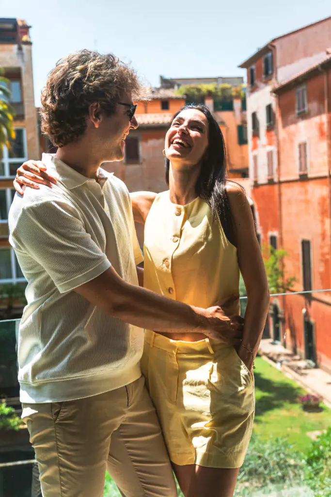 Happy couple smiling and embracing on a sunny balcony with colorful old buildings in the background.