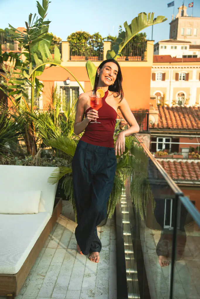 Woman enjoying a cocktail on a leafy rooftop terrace during golden hour in the city.